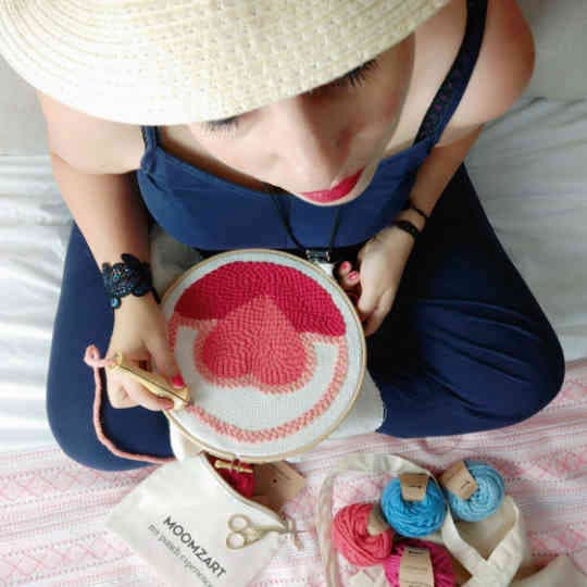 Woman embroidering with an Oxford needle on a wooden frame and merino wool, creating a heart design in punch needle.